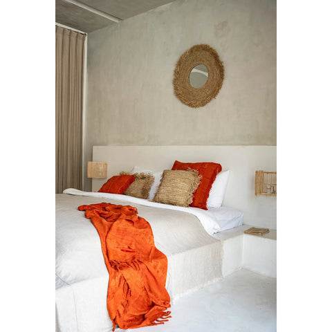 Bedroom with a bed featuring red and beige pillows and an orange throw, with a decorative mirror on the wall.