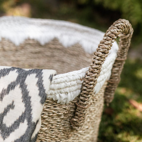 A close-up of the Round Seagrass Baskets WAIGEO with a sturdy handle, draped in a gray and white patterned cloth. The handmade storage basket rests on grass, surrounded by softly blurred greenery in the background.