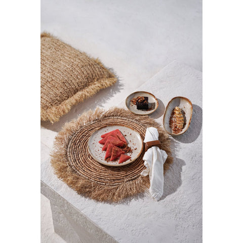 A plate with triangular slices of watermelon on handmade tableware rests on a woven placemat, next to two small dishes with desserts and a napkin on a white textured surface, with a fringed burlap cushion nearby.