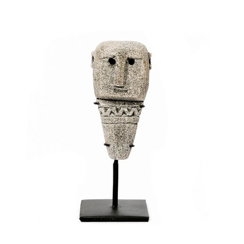 A stone sculpture resembling a simplified human figure, with a rectangular head, round eyes, a small nose, and a patterned midsection. Hand-carved by traditional Indonesian artisans, it is mounted on a black stand against a white background.