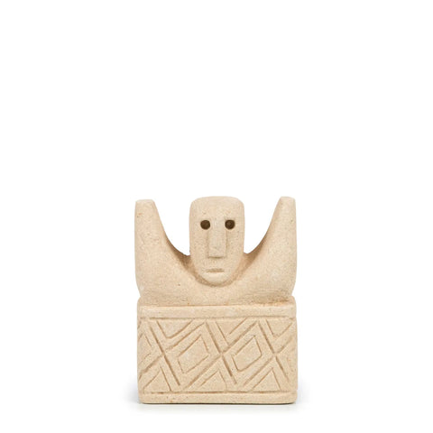 This small, beige sandstone sculpture from Sumba, Indonesia, features a stylized human figure with raised arms. The base is decorated with geometric patterns. The figure's simple, rounded face has minimal features, set against a plain white background.