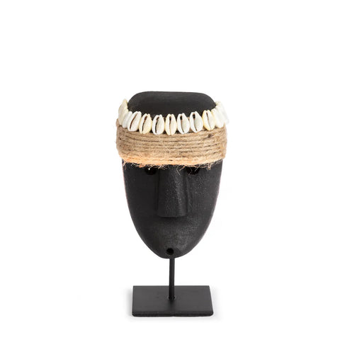 A black tribal mask on a stand, featuring a twine headband adorned with white cowrie shells, mimics the simplicity and craftsmanship of Sumba stone statues. The mask's carved facial features are set against a plain white background.