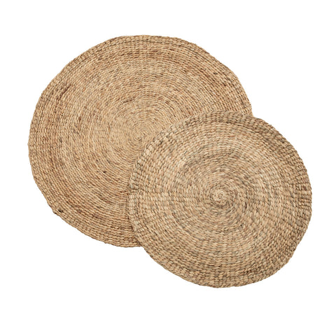 Two round, woven straw placemats are stacked, with the smaller one partially overlapping the larger. The texture is coarse and natural, reminiscent of water hyacinth weaving. Visible strands form concentric circles in a light, natural beige hue.