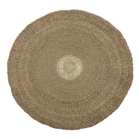 A round, woven jute rug with a natural beige color and a subtle gradient pattern radiating from the center to the edges. Its coarse, organic texture evokes a coastal feel, making it suitable for rustic or minimalist decor.