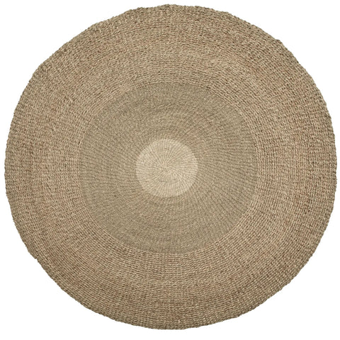 A large, round woven jute rug with a coastal feel, showcasing varying shades of natural beige. The texture appears rough and organic, with concentric circular patterns radiating from the center to the edges.