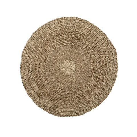 A round woven placemat made of natural fibers, displaying a spiral pattern from the center outward. Its coarse texture and natural beige color evoke the earthy tones of seagrass carpets, lending a subtle coastal feel reminiscent of the tranquil banks along the Mekong River.