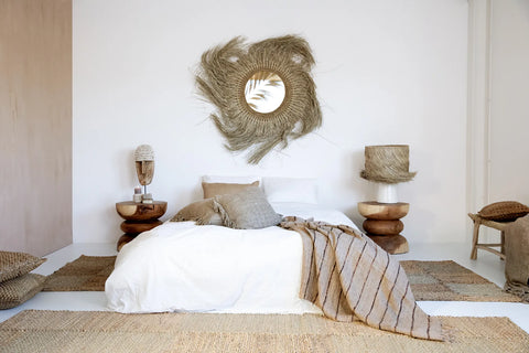 A minimalist bedroom features a white bed adorned with earthy-toned pillows and throws, complemented by a seagrass woven rug. Two natural wood side tables with unique lamps flank the bed, while a circular, textured wall mirror serves as the focal point above this serene space.