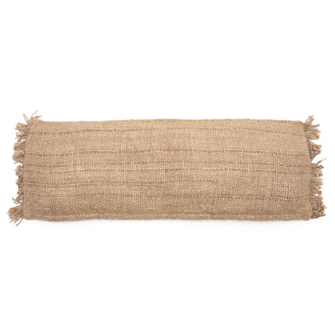 A cylindrical burlap pillow with frayed edges displays a textured, natural fiber surface. This boho cushion, laid horizontally against a white background, showcases its rustic, organic design with an artisanal touch.