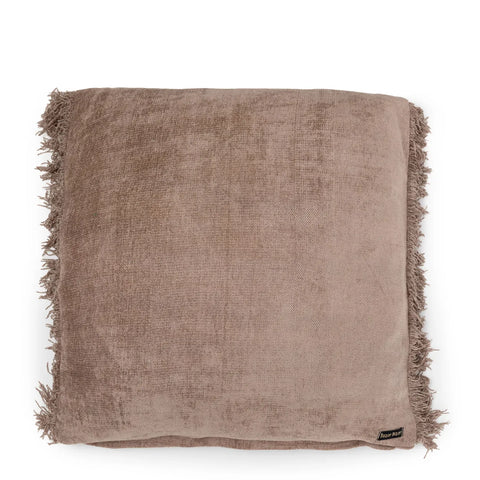 A square, light brown velvet cushion with fringed edges and a small black and beige tag on one corner embodies a hand-dyed, boho vibe. The cushion's soft and textured appearance adds a cozy feel.