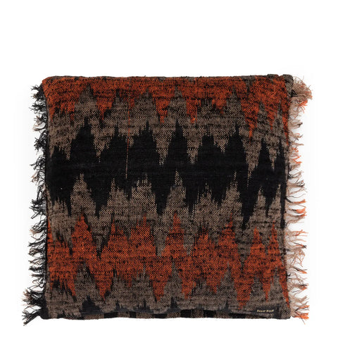 A square throw pillow exuding a boho vibe, featuring a geometric zigzag pattern. The design includes black and burnt orange hues on hand-dyed textile fabric with fringed edges.