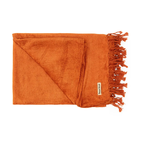 A folded, fringed orange throw blanket with a small fabric label sewn onto one corner. Its texture appears soft and plush, reminiscent of a luxury hand-woven plaid, making it ideal for cozying up.