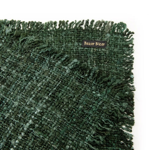 A close-up of a green woven rug with fringed edges boasts a hand-dyed look. A small rectangular label reads Bazar Bizar in light yellow text, attached to one corner of the rug. The texture is luxuriously coarse, and the color is a deep, forest green.