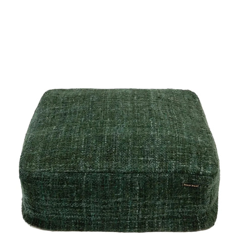 A square, forest green cotton pouffe with a textured fabric finish and a small tag on the side graces a plain white background. This hand-dyed home accessory brings an artisanal touch to any space.