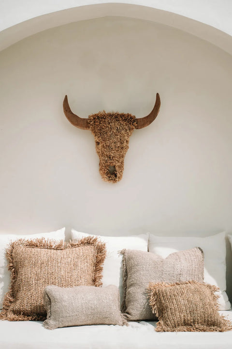 A white alcove features a decorative buffalo head wall hanging made of natural fibers, exuding a boho vibe. Below, a white couch is adorned with several hand-dyed cotton cushions in earthy tones, enhancing the cozy and rustic ambiance of the space.