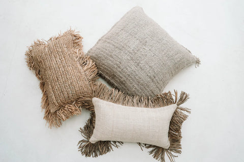 Three decorative pillows with fringe details, artfully arranged on a light surface. The collection includes a rectangular beige pillow, a square light grey cotton cushion, and an oval-shaped boho cushion with a woven texture in warm brown tones.