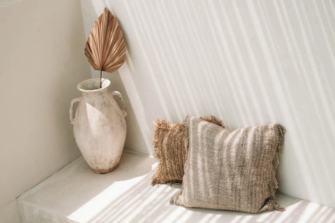 A minimalist scene featuring a large, textured white vase with a single dried palm leaf, placed on a ledge. Beside it, two hand-dyed cotton cushions rest against a white wall, casting gentle shadows from the slatted sunlight, adding to the boho vibe.