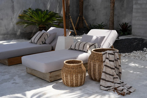 A serene outdoor seating area features two cushioned lounge chairs with boho cushions and striped pillows. Beside them are two woven baskets, one holding a matching blanket. The space is surrounded by lush plants and smooth stones against a concrete wall backdrop.