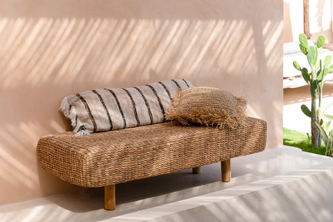 A woven rattan bench with wooden legs is adorned with a handmade textile and a fringed pillow. It sits on a light-colored patio, surrounded by sunlight casting striped shadows and a glimpse of greenery, creating a relaxed, boho vibe.