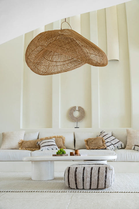 A minimalistic living room features a large, woven pendant light hanging over a white couch adorned with boho cushions. A low, round coffee table and a striped pouf sit in front of the couch, set against a backdrop of beige paneled walls.