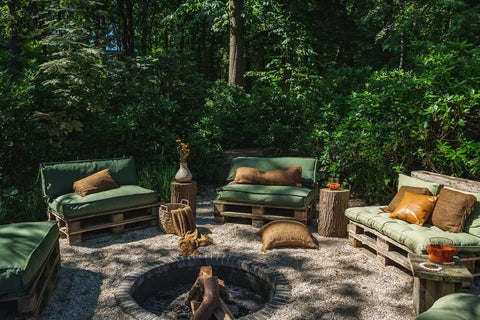 Garden lounge area with pallet furniture and boho vibe, featuring green and brown cotton cushions surrounding a circular stone fire pit. The setting is amidst dense green foliage and trees, with natural light filtering through the leaves, creating a rustic and inviting atmosphere.