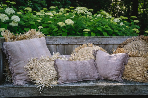 A rustic wooden bench adorned with a boho vibe features textured pillows in beige and lavender with fringed edges. A velvet cushion adds a touch of luxury, while lush green hydrangeas with white blooms flourish in the background.