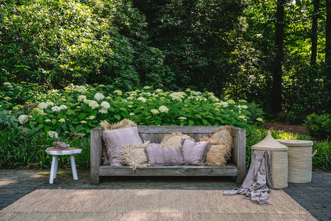 A rustic outdoor scene with a wooden bench adorned with boho vibe pillows and throws. Behind it, lush greenery and blooming white flowers create a serene backdrop. To the right, two woven baskets are placed beside the bench, and a small side table is on the left.