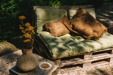 A cozy outdoor seating area showcases a rustic wood pallet sofa adorned with green cotton cushions and brown blankets. A vase with yellow flowers sits beside a boho cushion on a small wooden side table, all set against a gravel and garden backdrop.