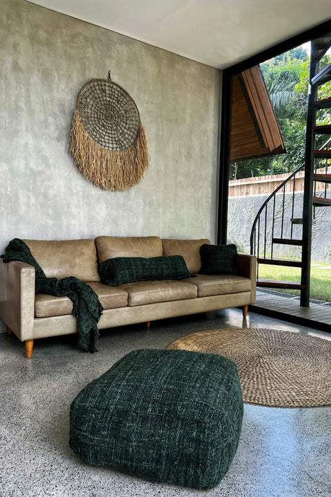 A modern living room features a beige sofa with green cushions, complemented by a hand-dyed cotton pouffe. A textured wall art piece hangs above, while a circular rug lies on the polished concrete floor. The spiral staircase near a large garden-view window adds an elegant touch to this chic home accessory ensemble.