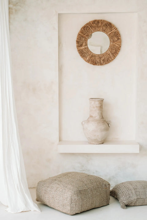 A minimalist room with a niche featuring a stone vase on a white shelf. Above the vase hangs a round mirror with a woven frame. Two textured cushions and a hand-dyed cotton pouffe lie on the floor nearby, while a sheer white curtain is partially visible on the left.