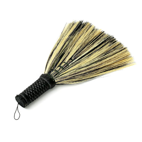 A small handheld whisk broom featuring a black woven handle and natural stiff bristles, ideal for light dusting or cleaning. Inspired by Indonesian hand sweepers, it adds a touch of Boho charm to any space.