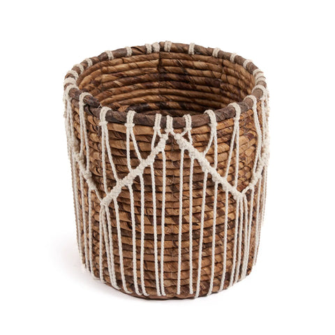 The Macra-Mazing Basket is a cylindrical, brown wicker creation wrapped with vertical white strings, creating a decorative macramé pattern. Its open top reveals a woven structure that complements the intricate design beautifully.