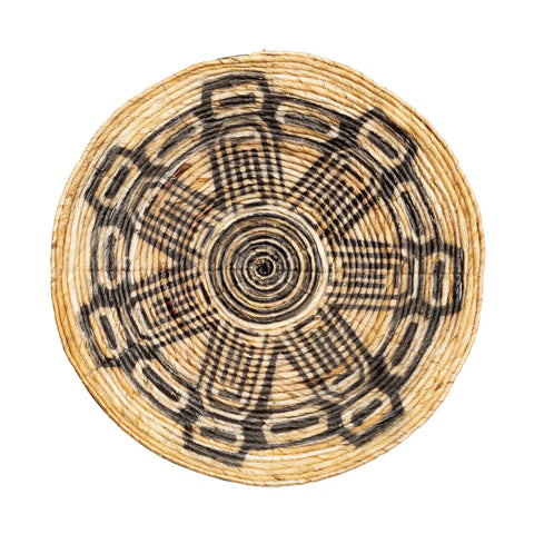 A handwoven plate with intricate black geometric patterns forms a star-like design, perfect for an ethnic wall display. Made of natural, light-colored fibers, this piece exudes a natural vibe.