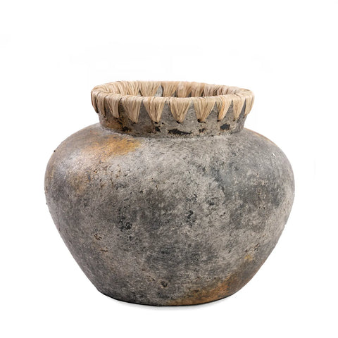 A rustic, weathered terracotta vase with a round body and wide mouth. The piece features a textured surface in shades of gray and brown, adorned with a decorative woven rim made of natural fibers, perfect for showcasing dried flowers.