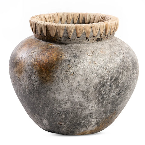 A rustic, antique grey clay pot with a textured surface and wide mouth. The rim is adorned with raffia detail, adding a natural touch. The pot shows signs of aging, with varied shades of gray and brown patches.