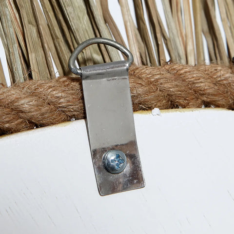 Close-up of a metal D-ring wall hanger attached to a wooden surface with a screw. The wood, partially covered by rope and straw-like materials, embodies an earthy boho vibe and suggests a natural mirror design with coastal textures.