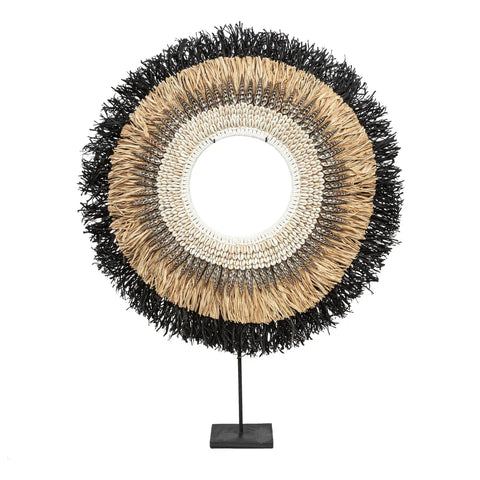 A circular woven decoration, this raffia hoop display includes a mirror at its center, featuring layers of beige, black, and cream-colored fibers. Hand-placed shells add a unique touch to this decorative item, which is elegantly mounted on a black stand.