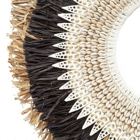 Close-up of a decorative item showcasing a circular pattern with layers of raffia display, white carved elements, and hand-placed shells. The colors transition from black to brown and white, creating a textured and intricate design.