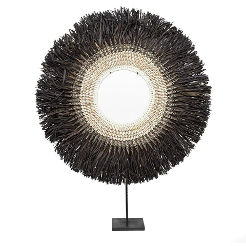 A decorative item with a circular design featuring a central round mirror, it is surrounded by layers of dark brown and beige textured materials resembling a sunburst pattern. This raffia display piece is mounted on a black stand, perfect for enhancing any home interior.