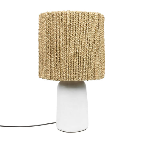 A boho lamp with a handwoven, textured beige shade rests on a smooth, white ceramic base. A black power cord extends from the base, completing the design that is both modern and minimalistic.