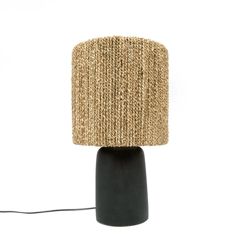 A bohemian table lamp featuring a handwoven straw lampshade and a matte black cylindrical base. A black cord extends from the base, resting on a white surface. This cozy lighting piece effortlessly combines rustic and modern elements.