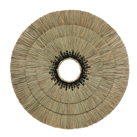 A round, handcrafted straw placemat showcases a radial pattern with layers fanning out from a central circle. Its textured appearance and earthy look make it an ideal bohemian coastal accent or wall deco piece.