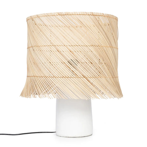 A rattan table lamp with a white cylindrical base and a beige, woven wicker lampshade offers a cozy light. The layered, textured design of the shade gives it a modern, rustic appearance. The lamp's power cord is subtly visible at the bottom left.