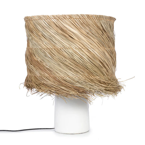 A pandan table lamp with a white cylindrical base and a large, woven straw shade in a rough, natural texture. The design resembles layers of dried grass, offering a boho look that is both rustic and organic. A cozy light glows warmly as the black electrical cord sits subtly at the base.