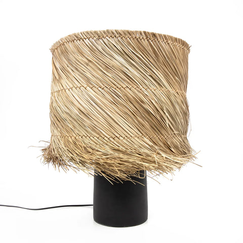 A pandan table lamp with a large woven straw lampshade, featuring fringed edges, adds a boho look. The cylindrical black base and visible electrical cord stand out against the plain white background.