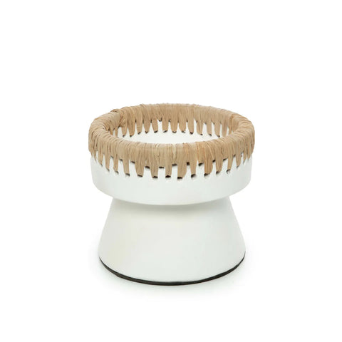 A small, white, round stool with a woven raffia seat and a wide, tapering base. The minimalist design showcases the natural texture of the material against the smooth, painted surface.