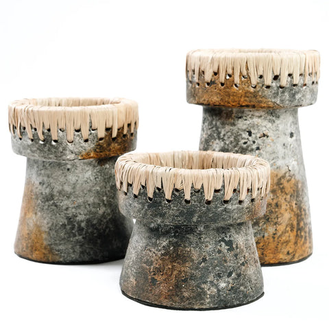 Three rustic stone planters of varying heights feature a cylindrical shape with woven wicker detailing around the top edge. The textured surface blends antique grey and terracotta tones, giving them a natural and earthy appearance, reminiscent of charming candle holders.