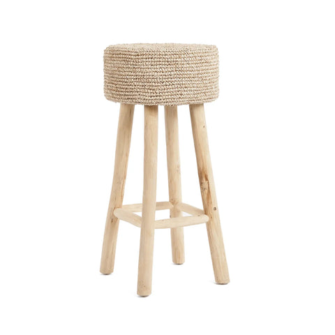 A raffia bar stool with four legs and a round, handwoven straw seat on top. Crafted from sustainable teak wood, it boasts a natural, rustic look against a plain white background.