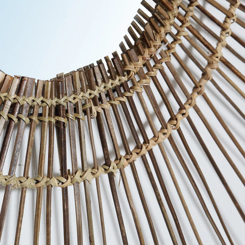 Close-up of a decorative item made from woven bamboo sticks arranged in a circular pattern. Resembling a 70s inspired mirror, the bamboo is tied with lighter fibers, crafting an intricate and textured design that evokes natural tropical decor against a light background.