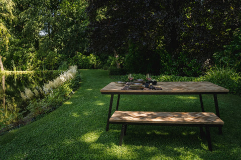 A teak wood picnic table with a bench is set with plates and glasses on a grassy area, beside a pond surrounded by lush green trees and bushes, creating a peaceful outdoor scene.