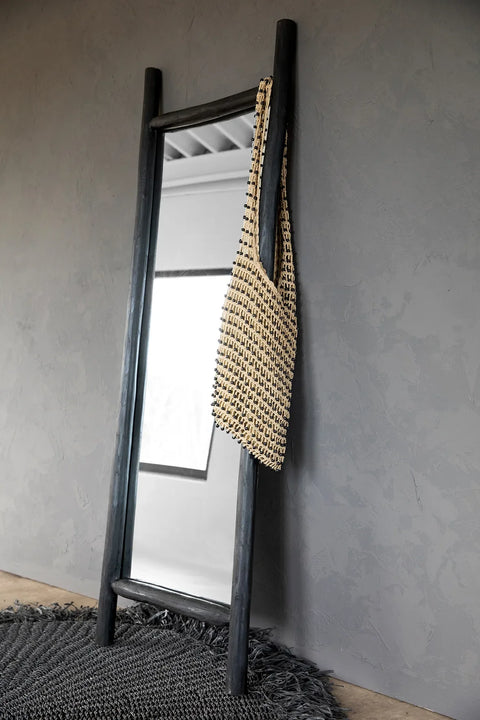 A tall, handcrafted teak ladder leans against a textured gray wall, adding a touch of decorative home interior charm. A woven tote bag hangs from one of the rungs, and the floor is carpeted with a dark, fringed rug. A reflective surface in the middle captures some interior light.